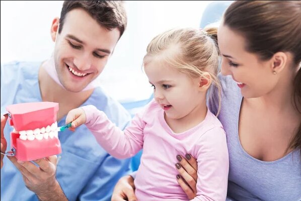 Medic stomatolog arătând unui copil cum se periază dinții, alături de mamă